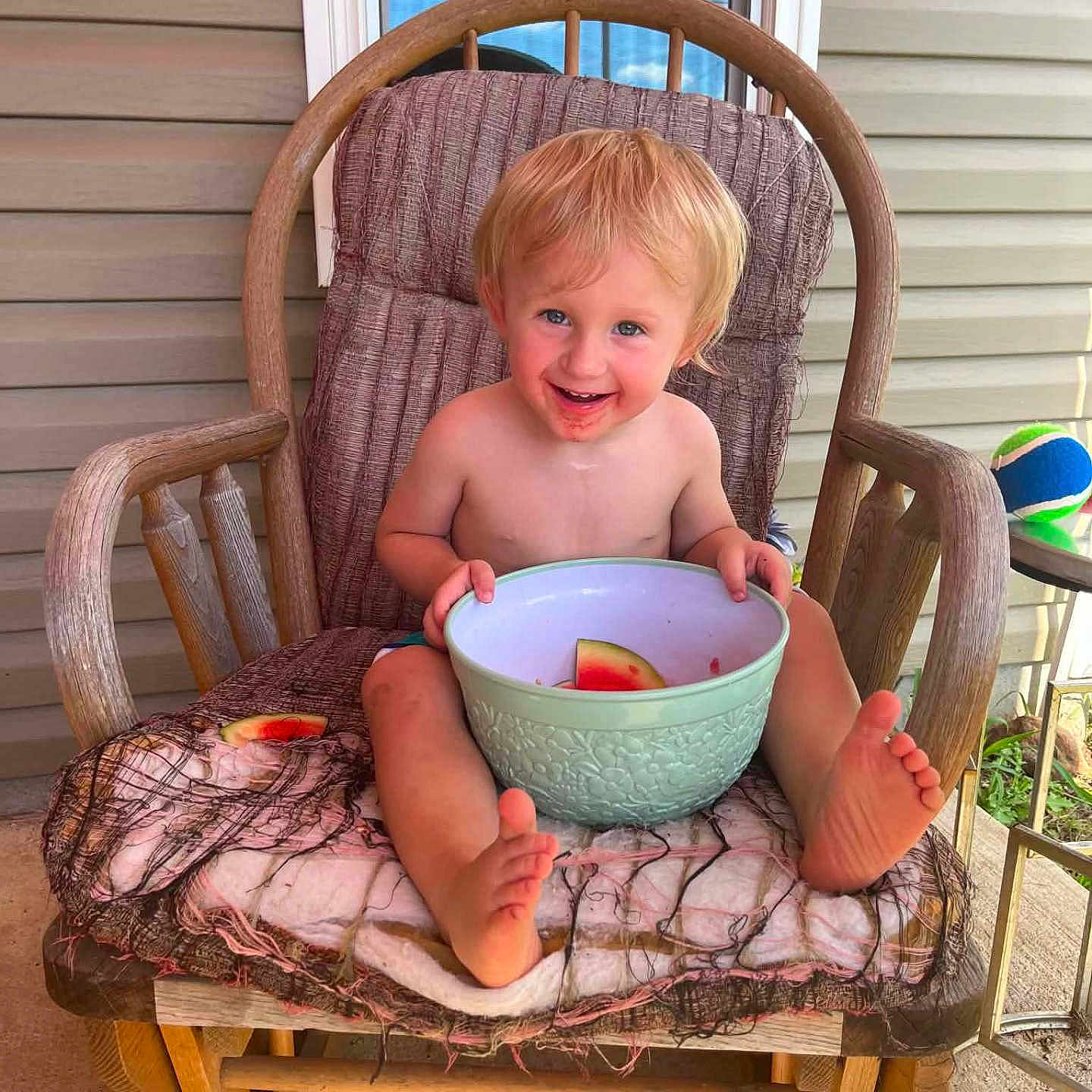 Liam is registered to the contest to win money with this photo: toddler, child, rocking_chair, watermelon, bowl, porch, window, blue_sky, ball, cushion, wooden_chair, smiling, barefoot, messy_face, summer, outdoor, happy, person, food, casual