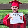 child, preschool, graduation, certificate, red_gown, red_cap, smile, outdoor, grass, fence, celebration, happy, person, young_child, achievement, portrait, event, daytime, school, success