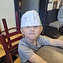 Konnor joined the competition — help win amazing prizes! child, paper_hat, drawing, animal_drawing, table, chair, indoor, person, blonde_hair, gray_shirt, seated, face, expression, playful, wall_art, furniture, wooden_chair, smile, kid, restaurant