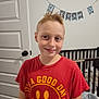 child, boy, smile, red_tshirt, short_hair, indoor, door, crib, banner, trash_bin, wood_floor, wall, home, casual_clothing, portrait, standing, happy, person, room, light