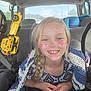 child, girl, smile, braid, face_paint, blanket, car_interior, seat, toy, yellow_toy, happy, casual_clothing, daylight, backseat, window, person, kid, candid, portrait, vehicle