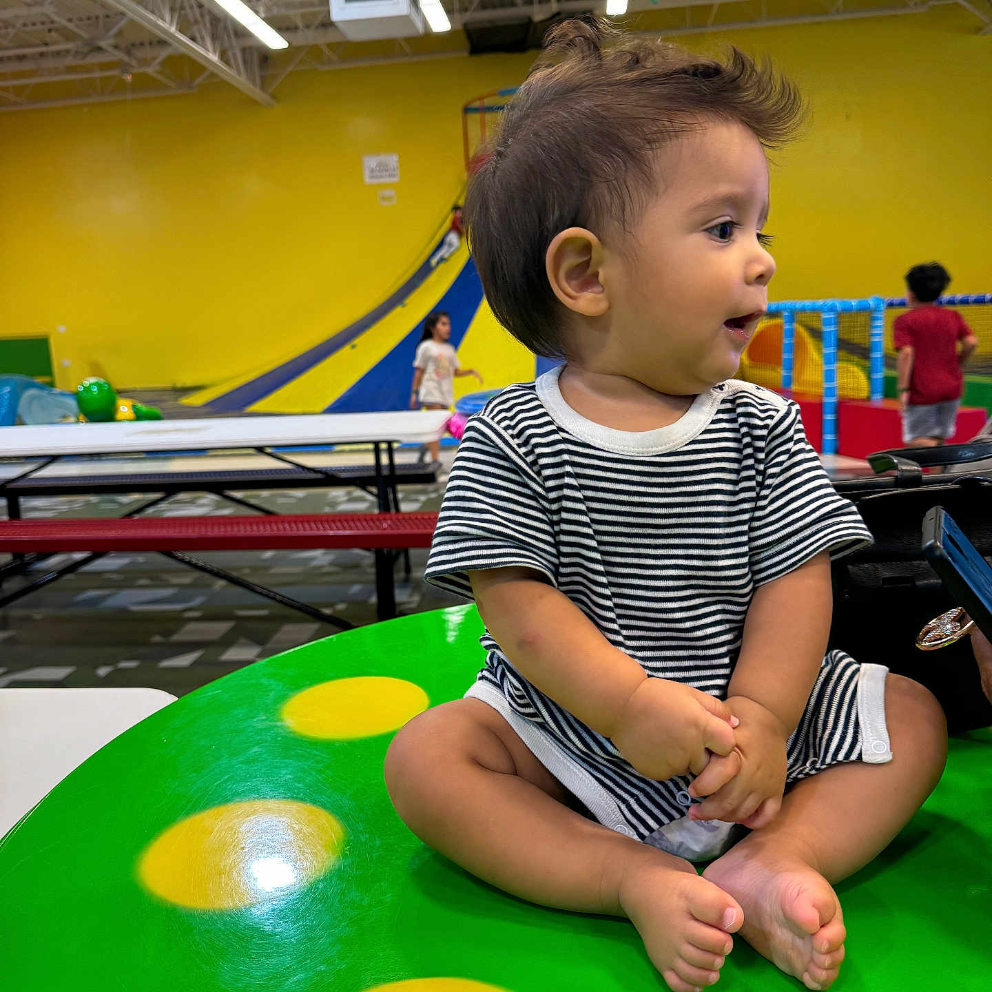 Amadeo joined the competition — help win amazing prizes! background, bench, child, colorful, cross_legged, curious, expression, floor, green_table, indoor, kids, play_area, playground, seated, short_hair, slide, striped_clothing, table, toddler, yellow_spots