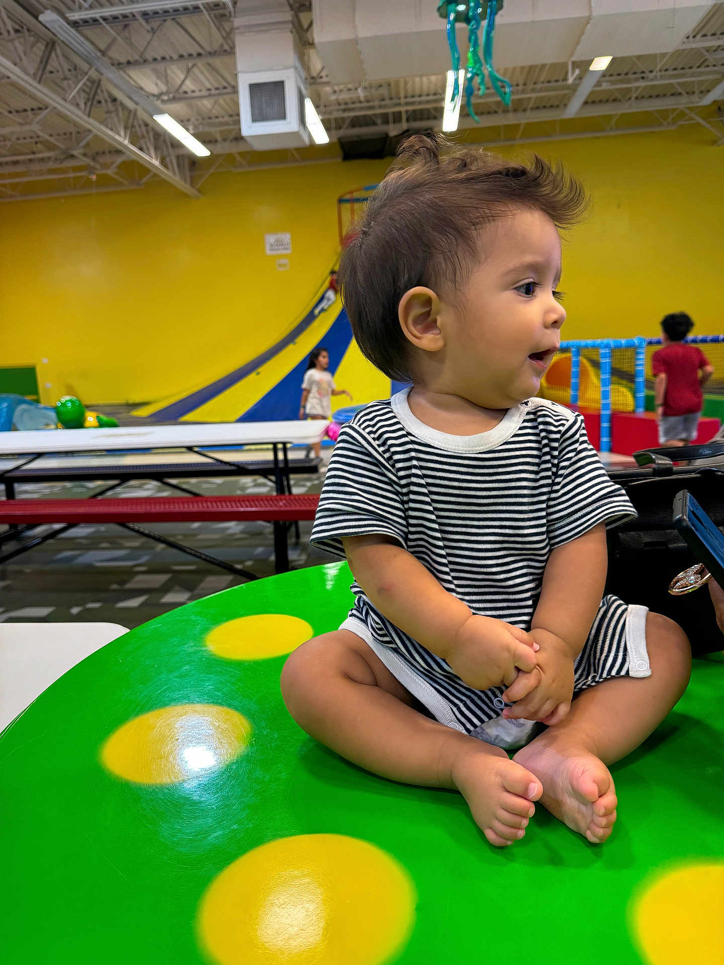 Amadeo joined the competition — help win amazing prizes! toddler, child, striped_clothing, indoor, play_area, table, green_table, yellow_spots, slide, kids, background, floor, bench, short_hair, curious, seated, cross_legged, expression, playground, colorful