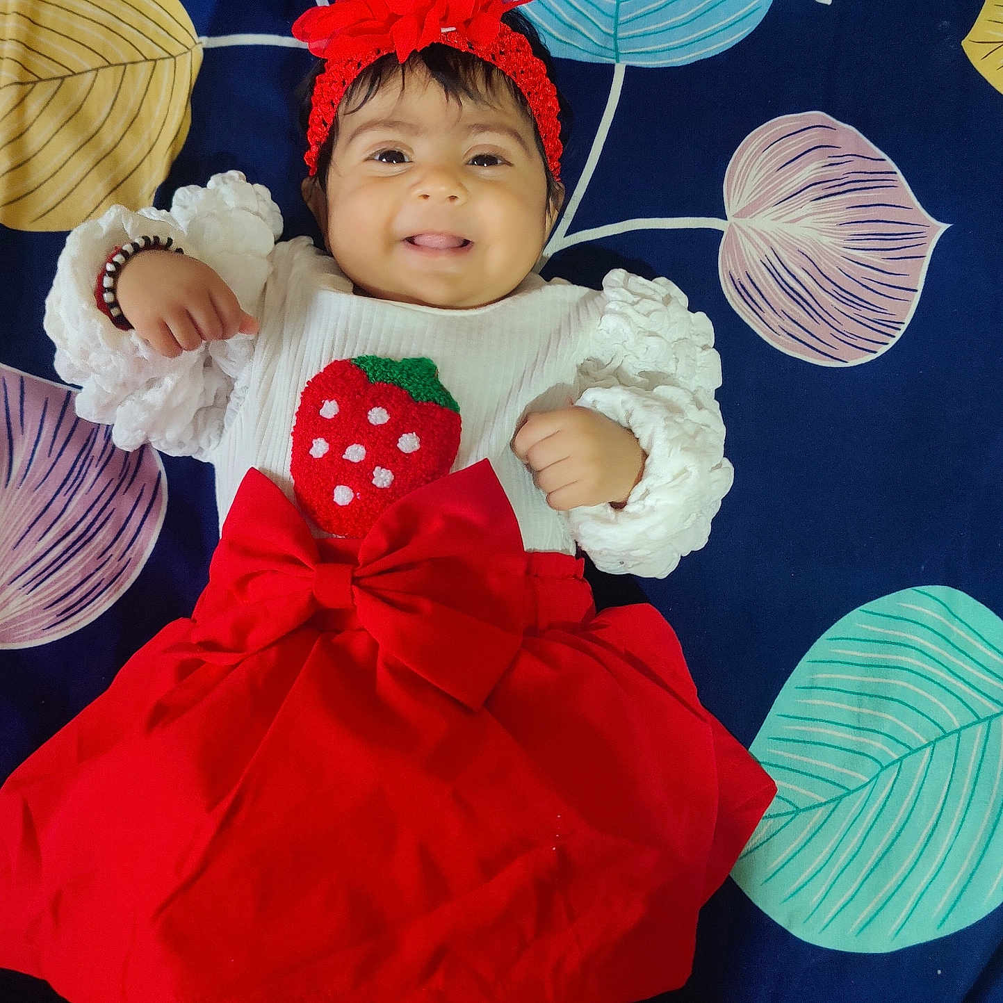 Nyra Adithi Darshan joined the competition — help win amazing prizes! baby, baby_girl, child, clothing, colorful_blanket, cute, fashion, flower_headband, happy, headband, indoors, infant, leaf_pattern, lying_down, person, portrait, red_skirt, smiling, strawberry_design, white_top