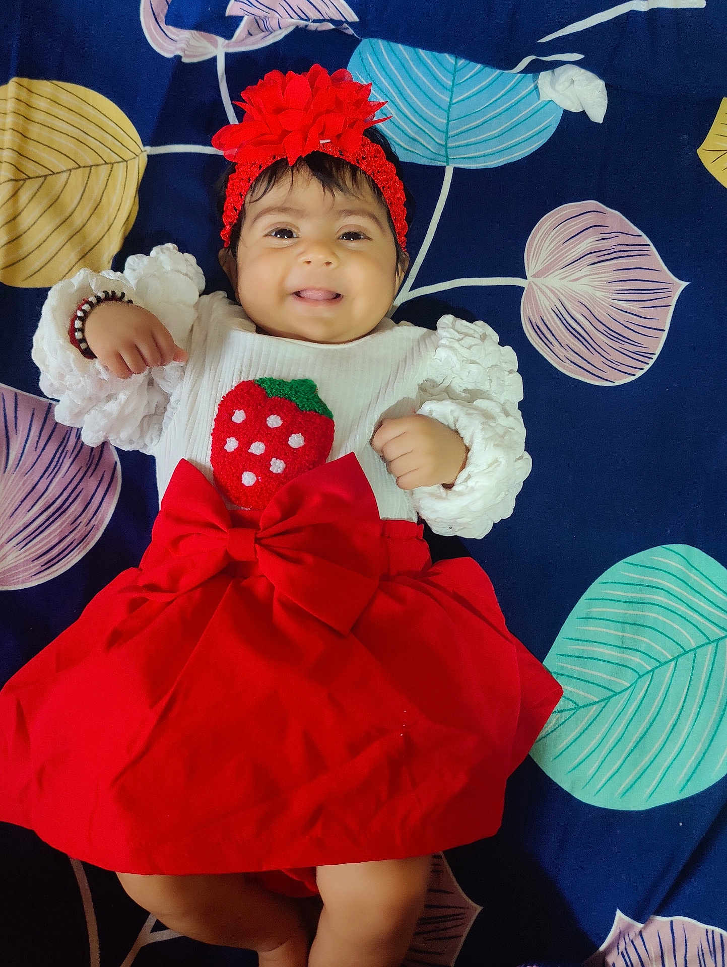 Nyra Adithi Darshan joined the competition — help win amazing prizes! baby, smiling, red_skirt, white_top, strawberry_design, headband, flower_headband, lying_down, colorful_blanket, leaf_pattern, cute, happy, infant, child, portrait, indoors, clothing, fashion, person, baby_girl