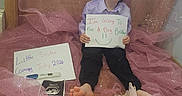 Roman is registered to the contest to win money with this photo: child, boy, smile, sign, announcement, pregnancy_test, ultrasound, pink_fabric, barefoot, baby_clothes, handwritten, sitting, indoor, window, curtain, happy, family, brother, sister, celebration