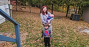 Roman is registered to the contest to win money with this photo: children, baby, toddler, mother, family, outdoor, autumn, fall_leaves, trees, yard, house, porch, grass, portrait, smiling, standing, baby_carrier, colorful_clothing, spiderman_shirt, family_photo