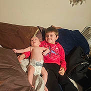 Roman joined the competition — help win amazing prizes! baby, child, toddler, siblings, portrait, smiling, sitting, lying_down, diaper, red_sweater, superhero_clothing, couch, pillows, blanket, indoor, bedroom, fan, feather_decoration, family, happy