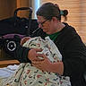 Chelsea is registered to the contest to win money with this photo: person, baby, newborn, blanket, footprint_pattern, glasses, hair_tie, indoor, stroller, bed, window_blinds, holding, cradling, seated, casual_clothing, adult, child, family, soft_lighting, portrait