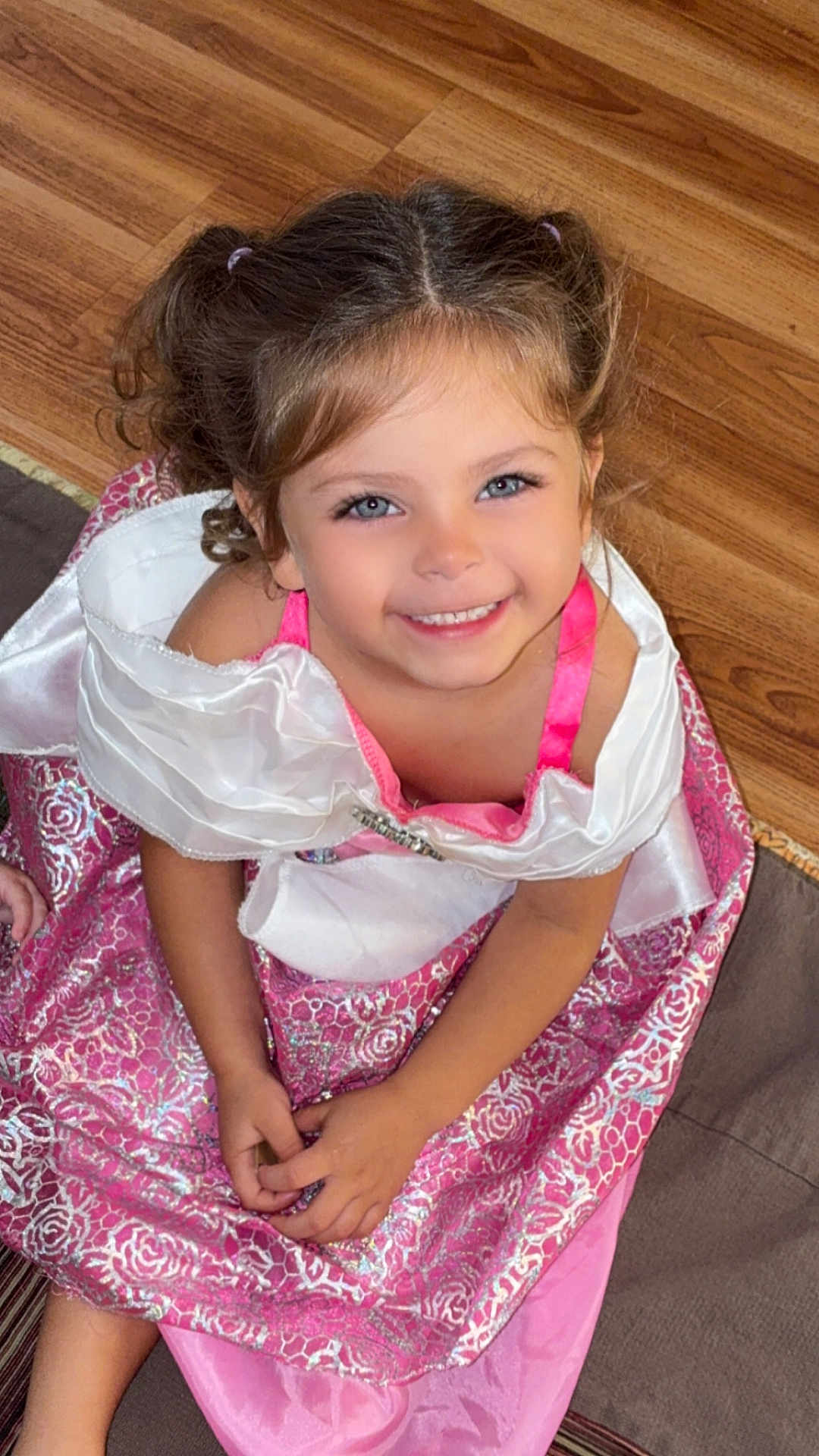 Honey is registered to the contest to win money with this photo: child, girl, smile, dress, princess, pink, white, curly_hair, pigtails, floor, wood, rug, happy, portrait, indoor, young, cute, sitting, hands, blue_eyes