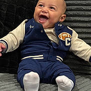 Phinesse is registered to the contest to win money with this photo: baby, infant, smiling, laughing, blue_jacket, white_socks, couch, plush, soft_texture, indoor, child, happy, cute, portrait, seated, clothing, face, expression, cozy, comfort