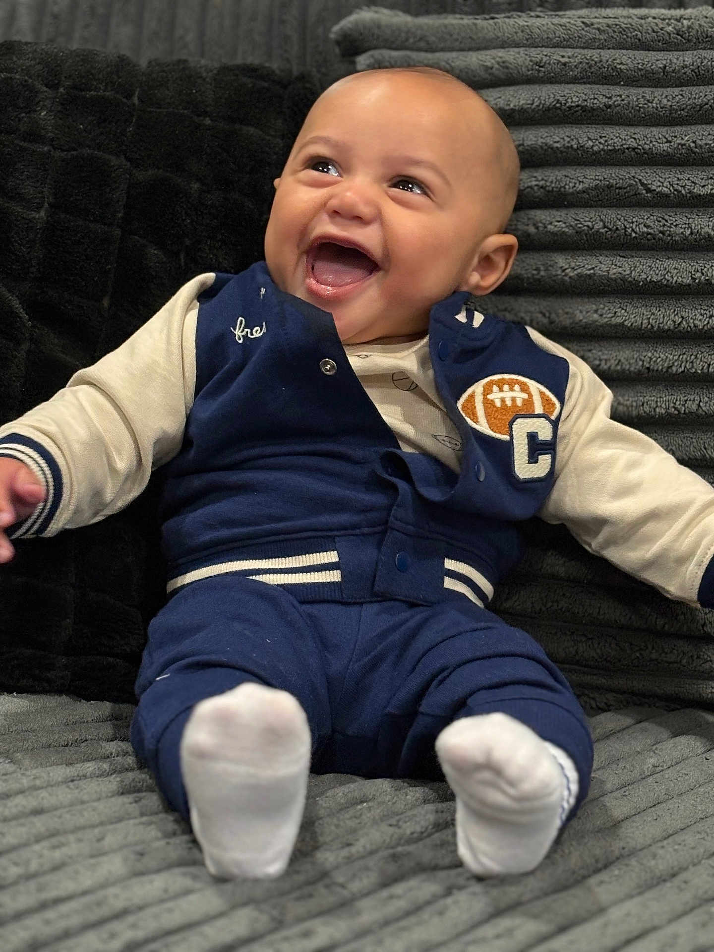Phinesse is registered to the contest to win money with this photo: baby, infant, smiling, laughing, blue_jacket, white_socks, couch, plush, soft_texture, indoor, child, happy, cute, portrait, seated, clothing, face, expression, cozy, comfort