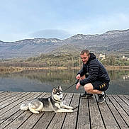 Aïka participe au concours pour gagner de l'argent avec cette photo : dog, husky, puppy, person, man, dock, wooden_boardwalk, lake, mountains, water_reflection, outdoors, leash, jacket, sneakers, sky, nature, pet_owner, portrait, relaxed, scenic_view