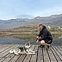 dog, husky, puppy, person, man, dock, wooden_boardwalk, lake, mountains, water_reflection, outdoors, leash, jacket, sneakers, sky, nature, pet_owner, portrait, relaxed, scenic_view