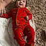 baby, blanket, carpet, child, cozy, cute, grinch, hand, happy, holiday_theme, indoor, infant, lying_down, pajamas, person, red_clothing, sleepwear, smiling, soft_texture, tattoo