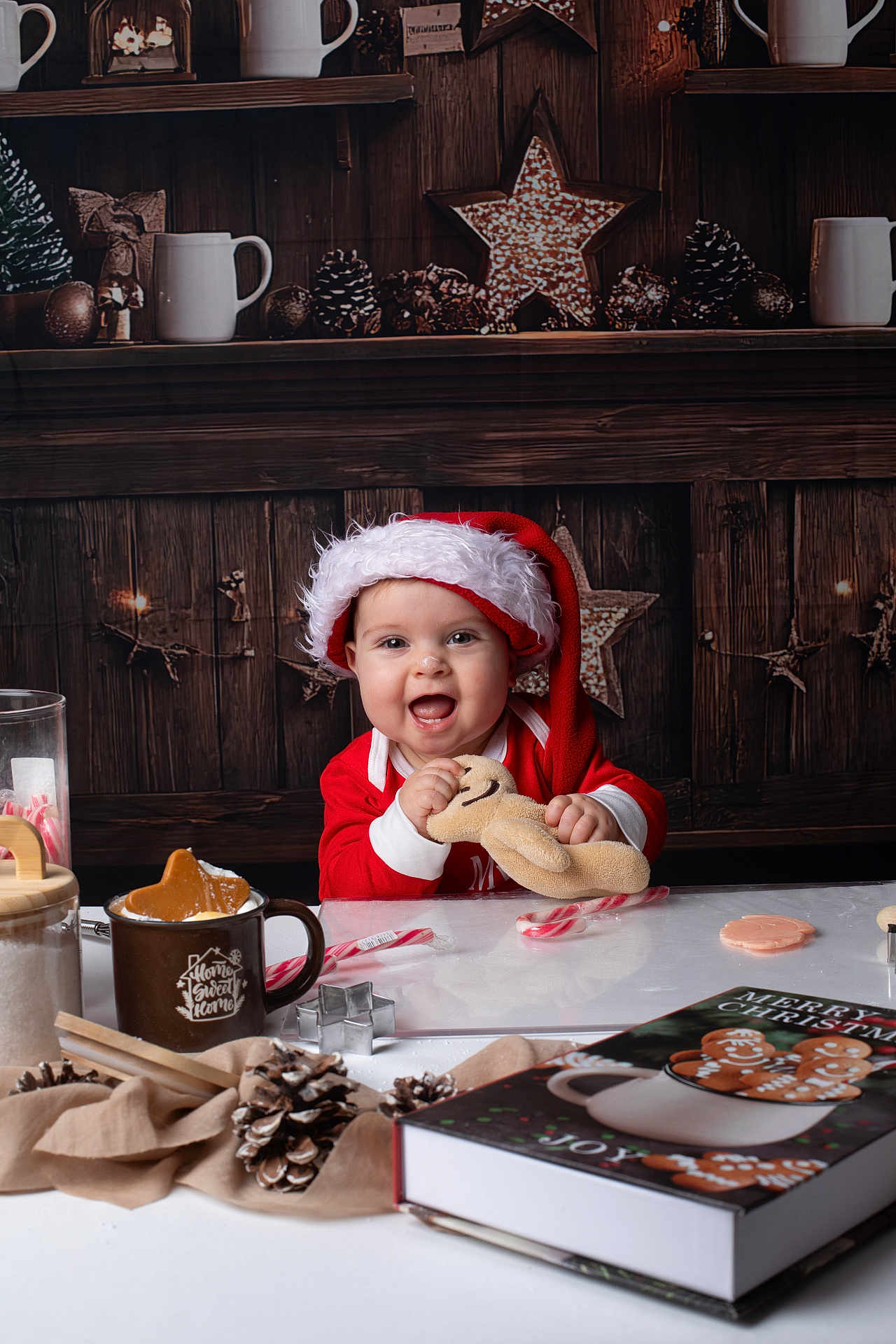 Eléa participe au concours pour gagner de l'argent avec cette photo : baby, child, santa_hat, red_clothing, gingerbread_plush, pinecones, candy_cane, cookie_cutter, mug, holiday_decor, christmas_book, wooden_background, table, smiling, festive, indoors, cute, toy, christmas_theme, happy