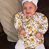 baby, child, infant, smiling, headband, white_tights, floral_outfit, couch, pillows, indoor, happy, person, seated, cute, headwear, toddler, fashion, comfort, soft_furniture, portrait