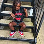 child, girl, sitting, stairs, outdoor, peace_sign, black_tshirt, cherry_graphic, denim_shorts, red_sneakers, bracelet, necklace, hair_bow, curly_hair, metal_stairs, concrete, greenery, tumbler, casual_clothing, playful