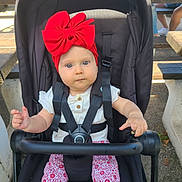 Agathe participe au concours pour gagner de l'argent avec cette photo : baby, child, stroller, red_bow, headband, outdoor, picnic_table, infant, seat_belt, white_shirt, patterned_pants, curious, sitting, daylight, people, casual, summer, park, bench, cute