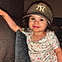 toddler, child, curly_hair, baseball_cap, smile, floral_shirt, pink_pants, couch, pillow, indoor, person, young_child, happy, casual_clothing, relaxed, cozy, home, portrait, cute, leaning