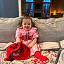 blanket, child, couch, cozy, curly_hair, curtains, furniture, happy, home_interior, indoor, living_room, person, pink_dress, red_belt, red_hat, red_socks, seated, smiling, toddler, window