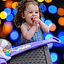 toddler, child, smiling, happy, curly_hair, toy, purple_toy, bokeh, colorful_lights, blue_lights, orange_lights, indoor, portrait, cute, playful, woven_surface, plastic_toy, baby_dress, clothing, young_child