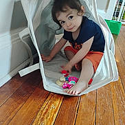 Ophelia joined the competition — help win amazing prizes! toddler, child, toy, basket, floor, wooden_floor, indoor, play, cute, person, young_child, sitting, colorful, curly_hair, casual_clothing, blue_shirt, orange_shorts, barefoot, home, fun