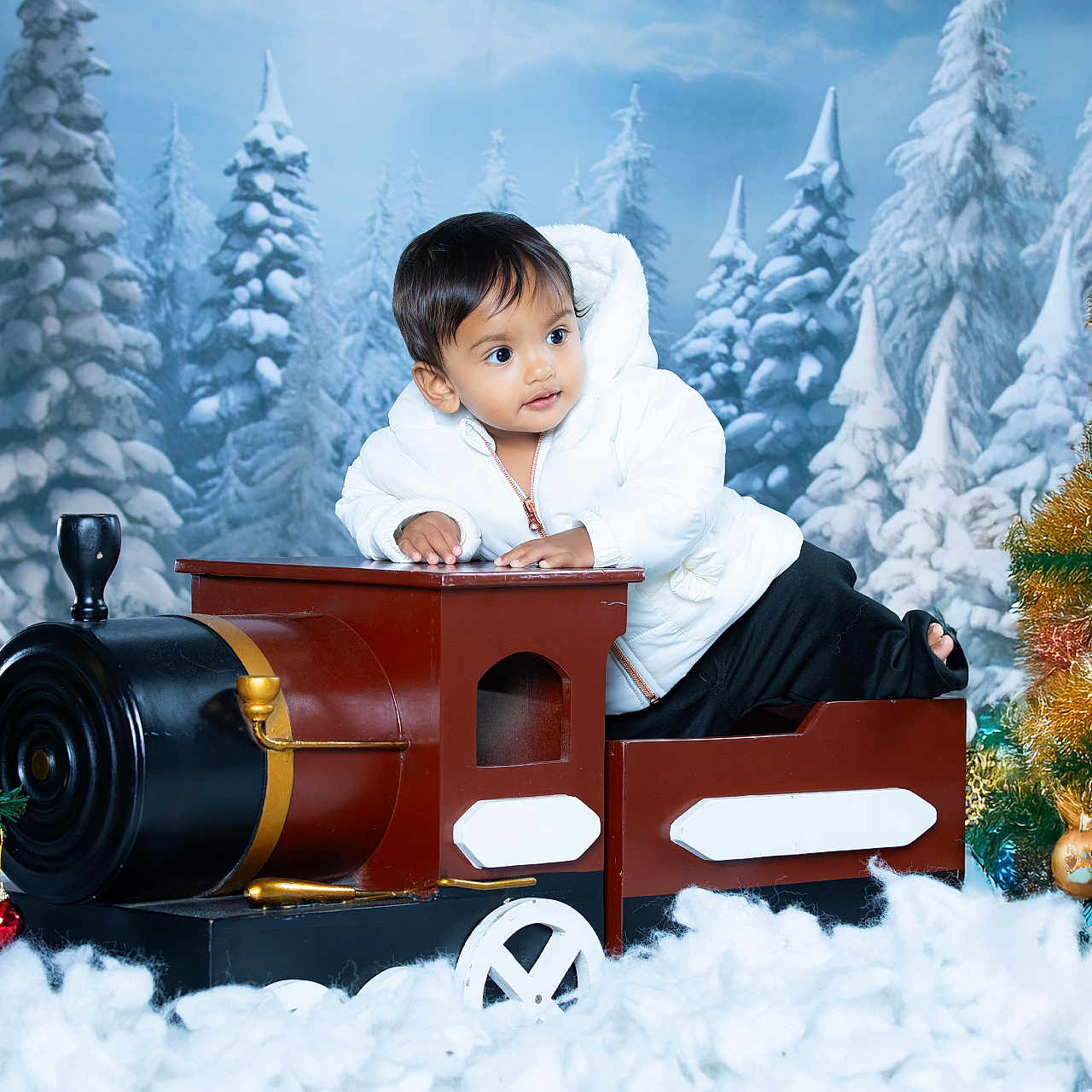 Varun is registered to the contest to win money with this photo: background, child, christmas_tree, curious, cute, decorations, festive, holiday, indoor, ornaments, play, portrait, seasonal, smiling, snow, toddler, toy_train, white_jacket, winter, winter_clothing