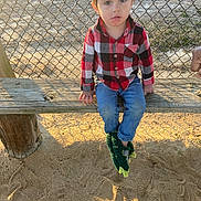 Preston is registered to the contest to win money with this photo: child, plaid_shirt, blue_jeans, green_slippers, wooden_bench, chain_link_fence, playground, sand, sunlight, outdoor, person, casual_clothing, dinosaur_slippers, young_child, seated, curious_expression, daylight, shadow, nature, recreation