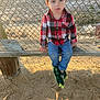 blue_jeans, casual_clothing, chain_link_fence, child, curious_expression, daylight, dinosaur_slippers, green_slippers, nature, outdoor, person, plaid_shirt, playground, recreation, sand, seated, shadow, sunlight, wooden_bench, young_child
