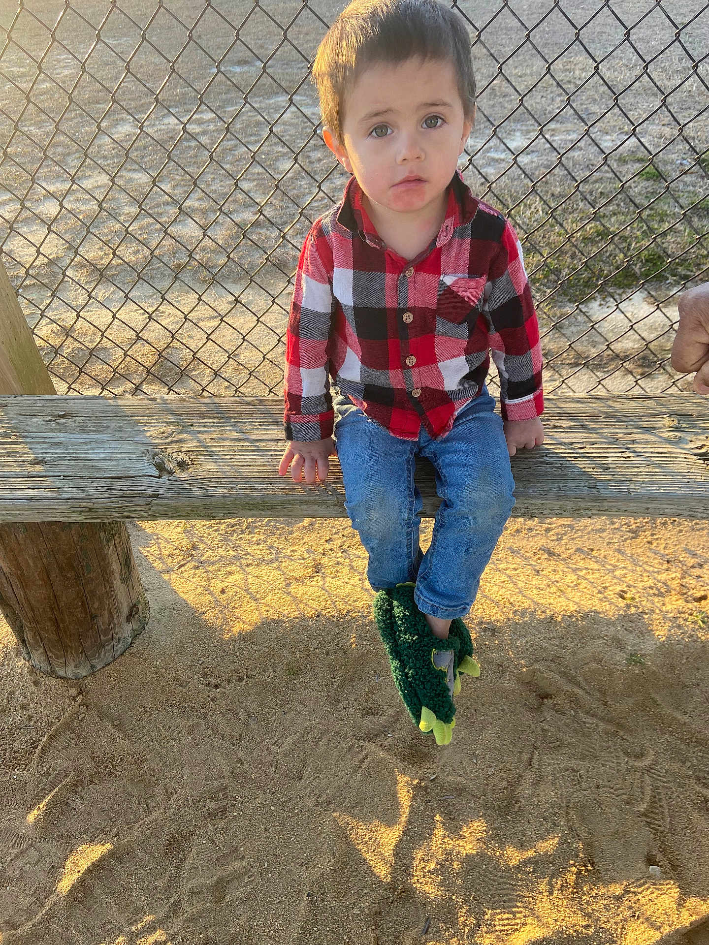 Preston is registered to the contest to win money with this photo: child, plaid_shirt, blue_jeans, green_slippers, wooden_bench, chain_link_fence, playground, sand, sunlight, outdoor, person, casual_clothing, dinosaur_slippers, young_child, seated, curious_expression, daylight, shadow, nature, recreation
