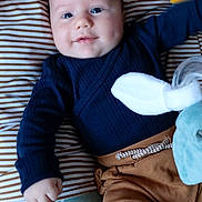 Jules participe au concours pour gagner de l'argent avec cette photo : baby, infant, smiling, face, cushion, striped, clothing, navy_blue, brown_pants, plush_toy, hand, indoor, soft_toys, blanket, cute, child, portrait, cozy, laying_down, comfort