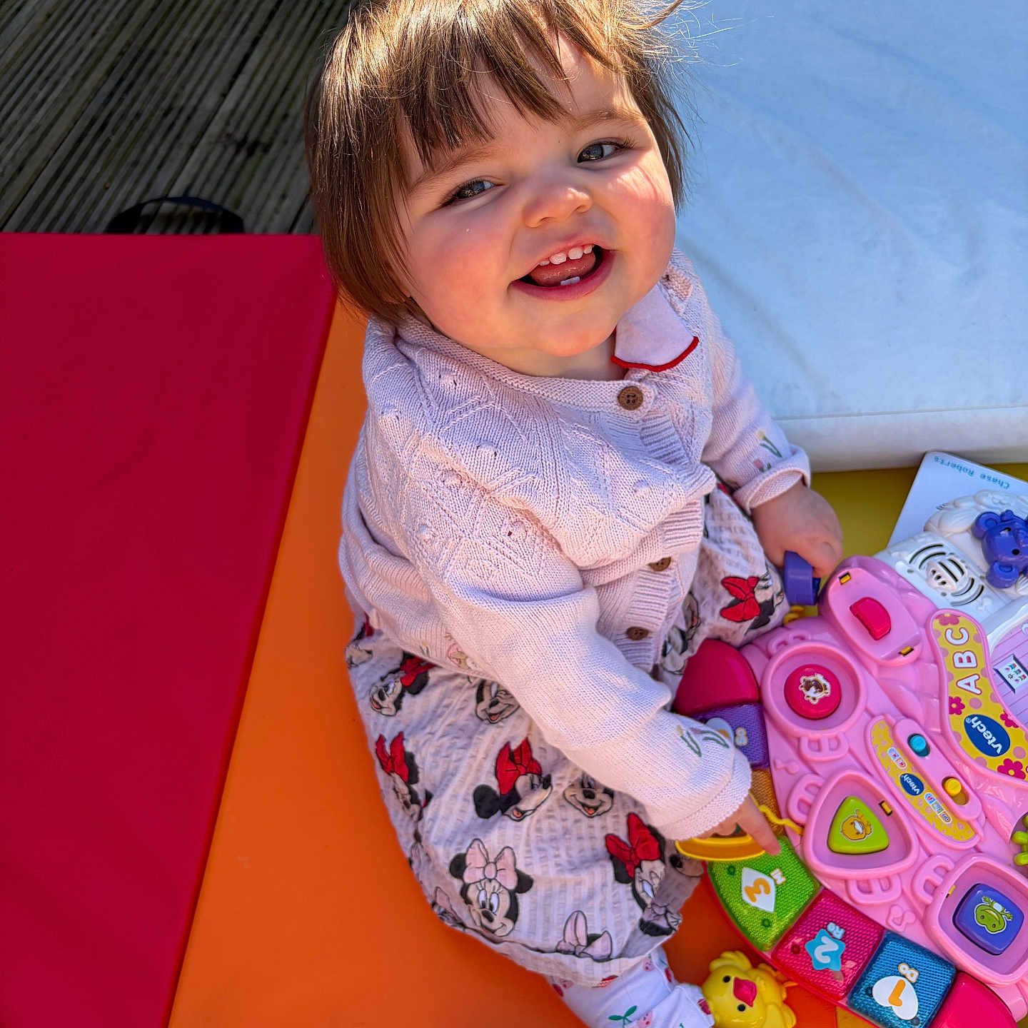 Elsie is registered to the contest to win money with this photo: toddler, child, smiling, toy, pink_toy, colorful, playtime, happy, cute, indoors, sitting, clothing, cardigan, patterned_pants, barefoot, sunlight, flooring, playmat, person, portrait