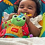 baby, child, plush_toy, frog_toy, blanket, baby_seat, smiling, indoors, curly_hair, person, toy, couch, robotic_vacuum, colorful, white_clothing, soft_texture, comfort, happy, infant, playtime