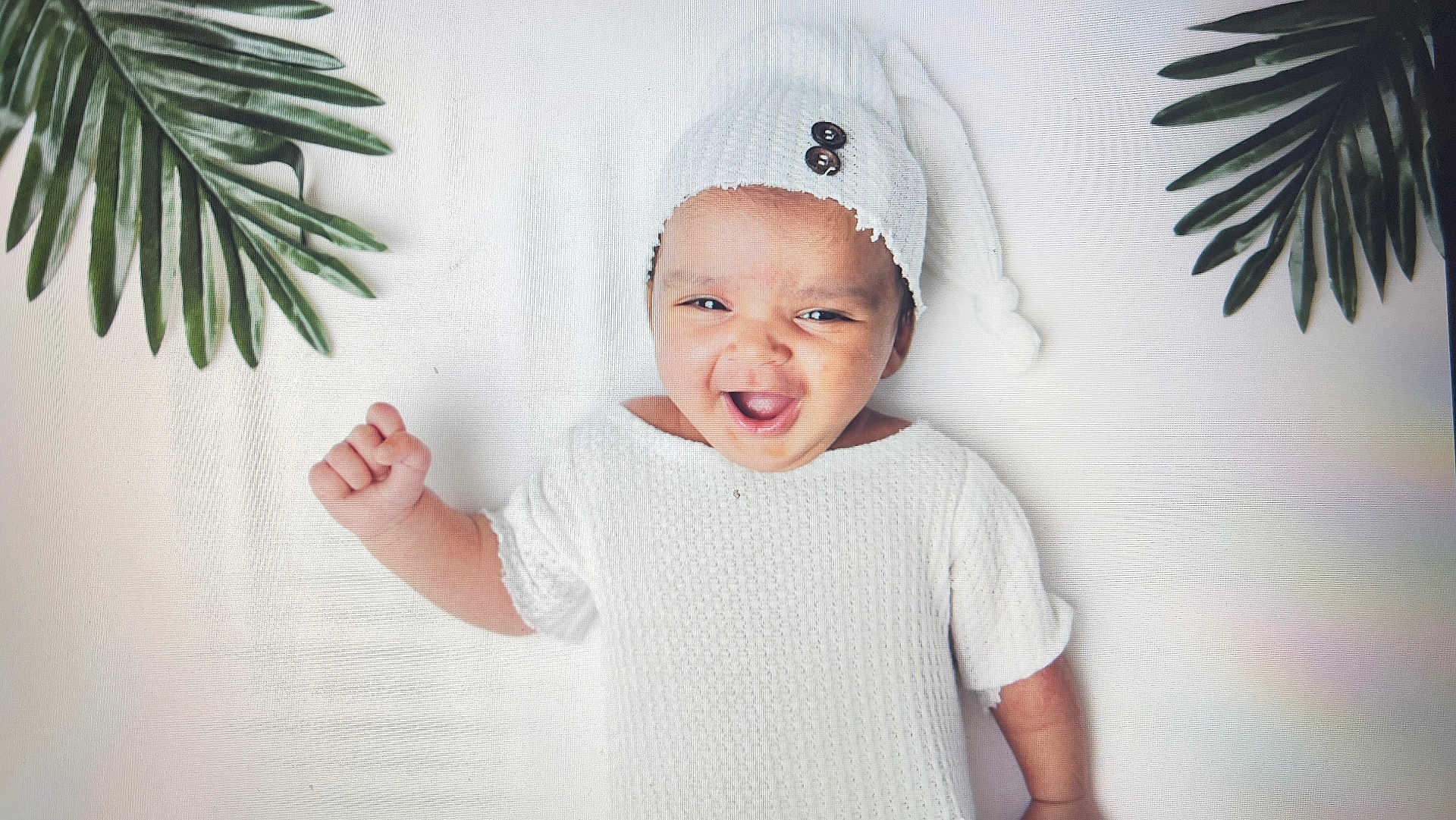 Pahal joined the competition — help win amazing prizes! baby, infant, smile, white_clothing, knit_hat, raised_fist, green_leaves, happy, portrait, cute, child, indoors, soft_background, button_detail, closeup, baby_gesture, cheerful, lying_down, adorable, young