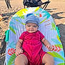 baby, infant, child, cap, red_clothing, baby_seat, beach, sand, outdoor, sunlight, trees, chair, blue_sky, person, footwear, socks, summer, recreation, relaxation, nature