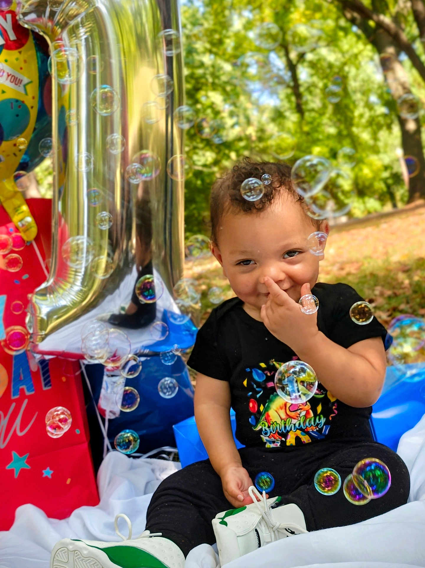 Zaiden is registered to the contest to win money with this photo: toddler, child, birthday, balloon, bubbles, outdoor, park, gift_bags, smiling, black_clothing, white_shoes, curly_hair, playful, celebration, happy, sunlight, greenery, sitting, face, cute
