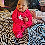 Zaiden is registered to the contest to win money with this photo: baby, child, red_clothing, onesie, smiling, sitting, bed, zebra_print, blanket, headboard, string_lights, cozy, indoor, portrait, happy, person, furniture, sleepwear, cute, infant