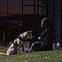 Canela is registered to the contest to win money with this photo: dog, person, grass, fence, dusk, sunset, outdoor, leash, harness, sitting, glasses, casual_clothing, side_profile, evening, shadow, building, quiet, contemplative, friendship, relaxing