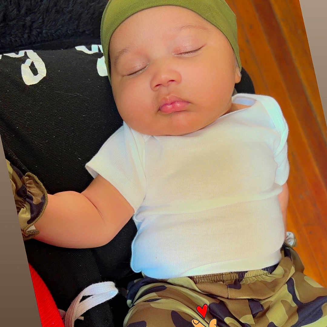 Chozien is registered to the contest to win money with this photo: baby, sleeping, infant, green_cap, white_shirt, camo_pants, cushion, black_cushion, floor, wood_floor, cute, peaceful, resting, indoor, baby_clothing, sock, hand, emoji_overlay, heart, closeup