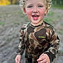 child, toddler, curly_hair, blue_eyes, smiling, camouflage_clothing, outdoor, portrait, happy, face, long_sleeve_shirt, nature, young_child, person, daylight, casual_wear, standing, skin, hand, grass