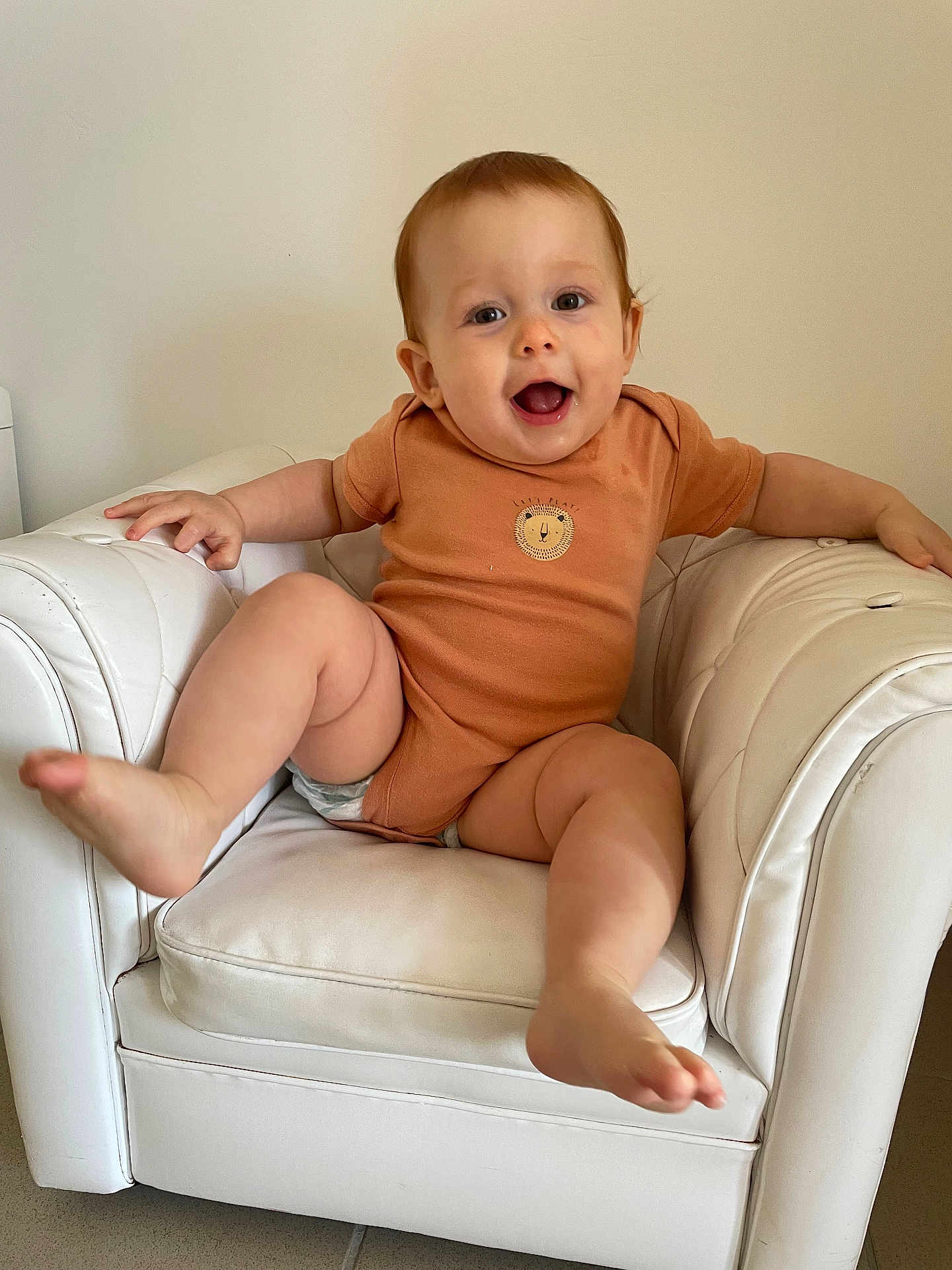 Sylvia Maria a rejoint le concours — aidez-le/la à gagner de superbes lots ! baby, casual_clothing, child, cute, floor_tiles, furniture, happy, home, indoor, infant, kicking_leg, leather_chair, lion_emblem, onesie, person, playful, seated, smiling, white_chair, young_child