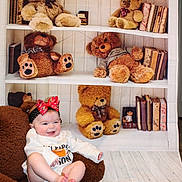 Raylynn is registered to the contest to win money with this photo: baby, child, smiling, bow, headband, teddy_bear, chair, bookshelf, books, toy, floor, indoor, clothing, happy, infant, portrait, seated, soft_texture, background, cute