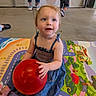 background_people, blue_dress, casual, child, colorful, concrete_floor, curly_hair, feet, floor, indoor, legs, person, play_mat, playtime, red_ball, sitting, smiling, toddler, toy, young_child
