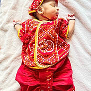 Leyan participe au concours pour gagner de l'argent avec cette photo : baby, sleeping, red_clothing, traditional_clothing, headwear, blanket, infant, cute, child, soft_texture, indoors, resting, peaceful, small_hands, footwear, patterned_fabric, portrait, cultural, newborn, lying_down