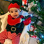 baby, child, christmas_elf_costume, christmas_tree, holiday, festive, gift_wrap, smiling, indoor, toy, decorations, red_and_green, striped_pants, hat, blanket, floor, face, joyful, holiday_season, celebration