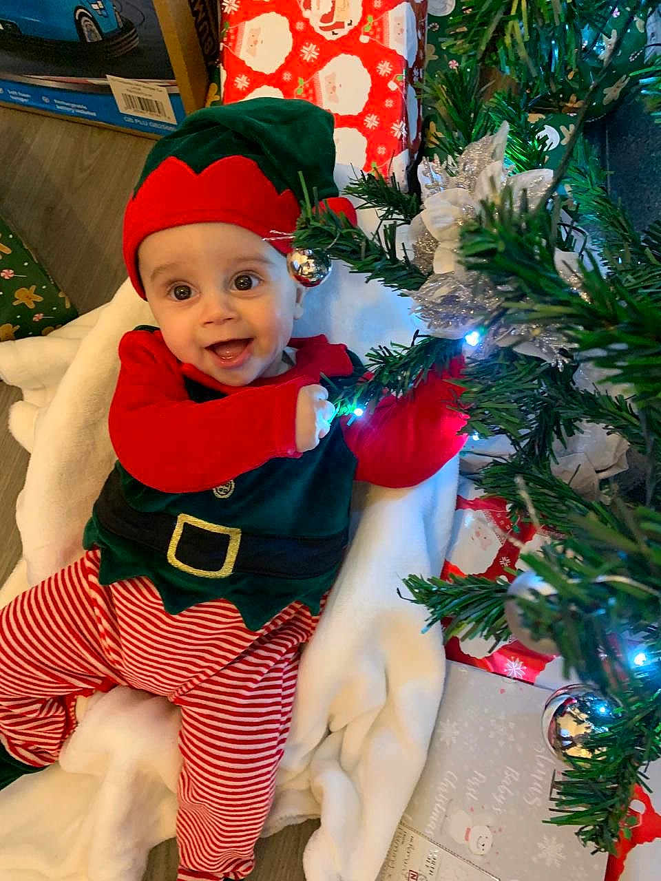 Roman is registered to the contest to win money with this photo: baby, child, christmas_elf_costume, christmas_tree, holiday, festive, gift_wrap, smiling, indoor, toy, decorations, red_and_green, striped_pants, hat, blanket, floor, face, joyful, holiday_season, celebration