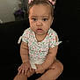 baby, child, girl, sitting, floral_shirt, shorts, pink_bows, hair_accessory, indoors, couch, black_background, curious_expression, skin, face, hands, legs, casual_clothing, portrait, young_child, cute
