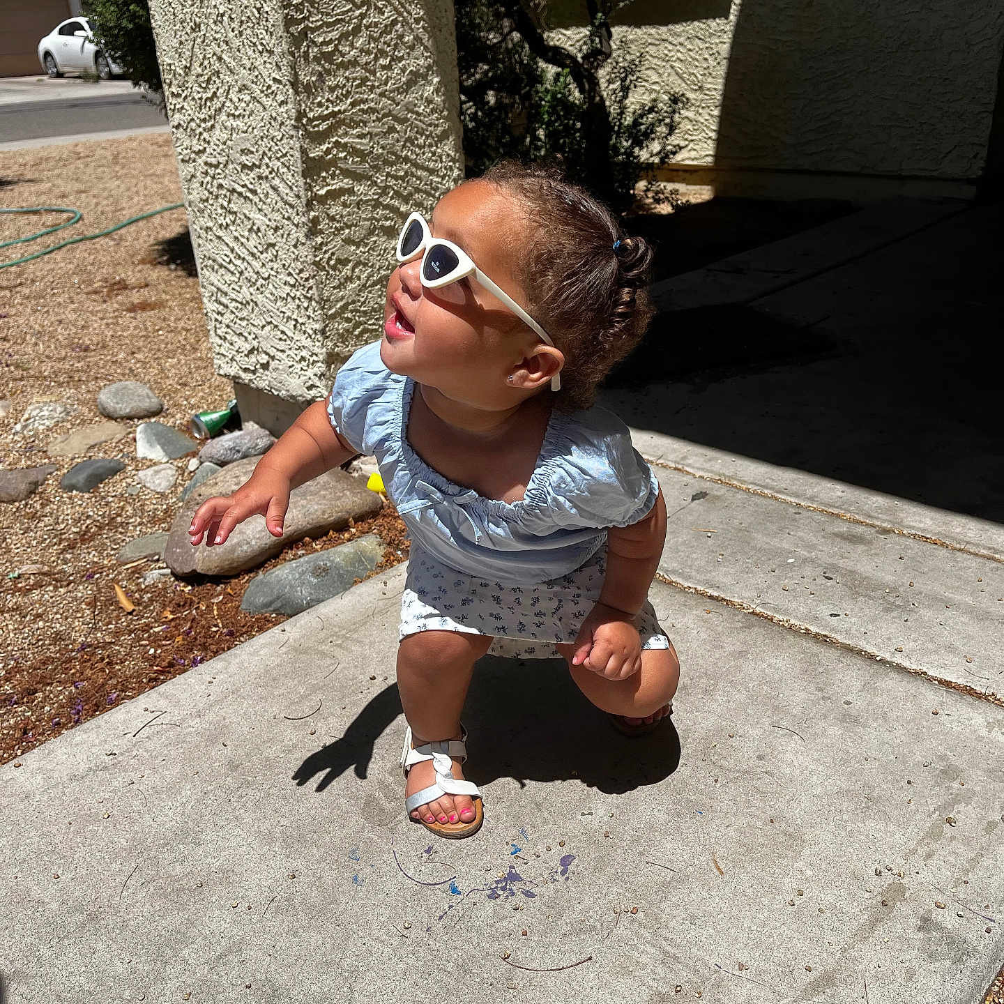 Rayanna is registered to the contest to win money with this photo: building, child, concrete, crouching, curly_hair, dress, outdoor, plants, playful, residential_area, rocks, sandals, shadow, sidewalk, smiling, summer, sunglasses, sunlight, toddler, window