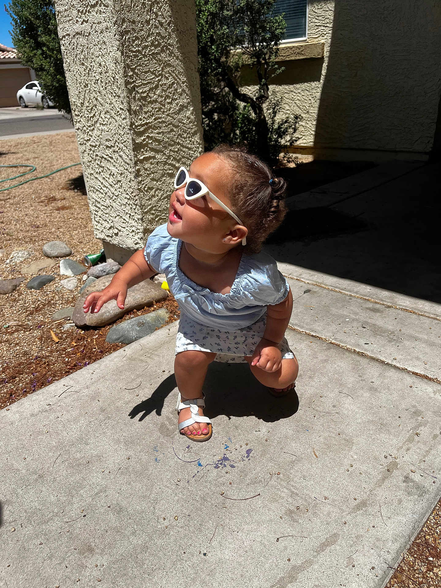Rayanna is registered to the contest to win money with this photo: toddler, child, sunglasses, outdoor, sidewalk, sunlight, shadow, concrete, dress, sandals, curly_hair, playful, summer, smiling, crouching, building, window, rocks, plants, residential_area
